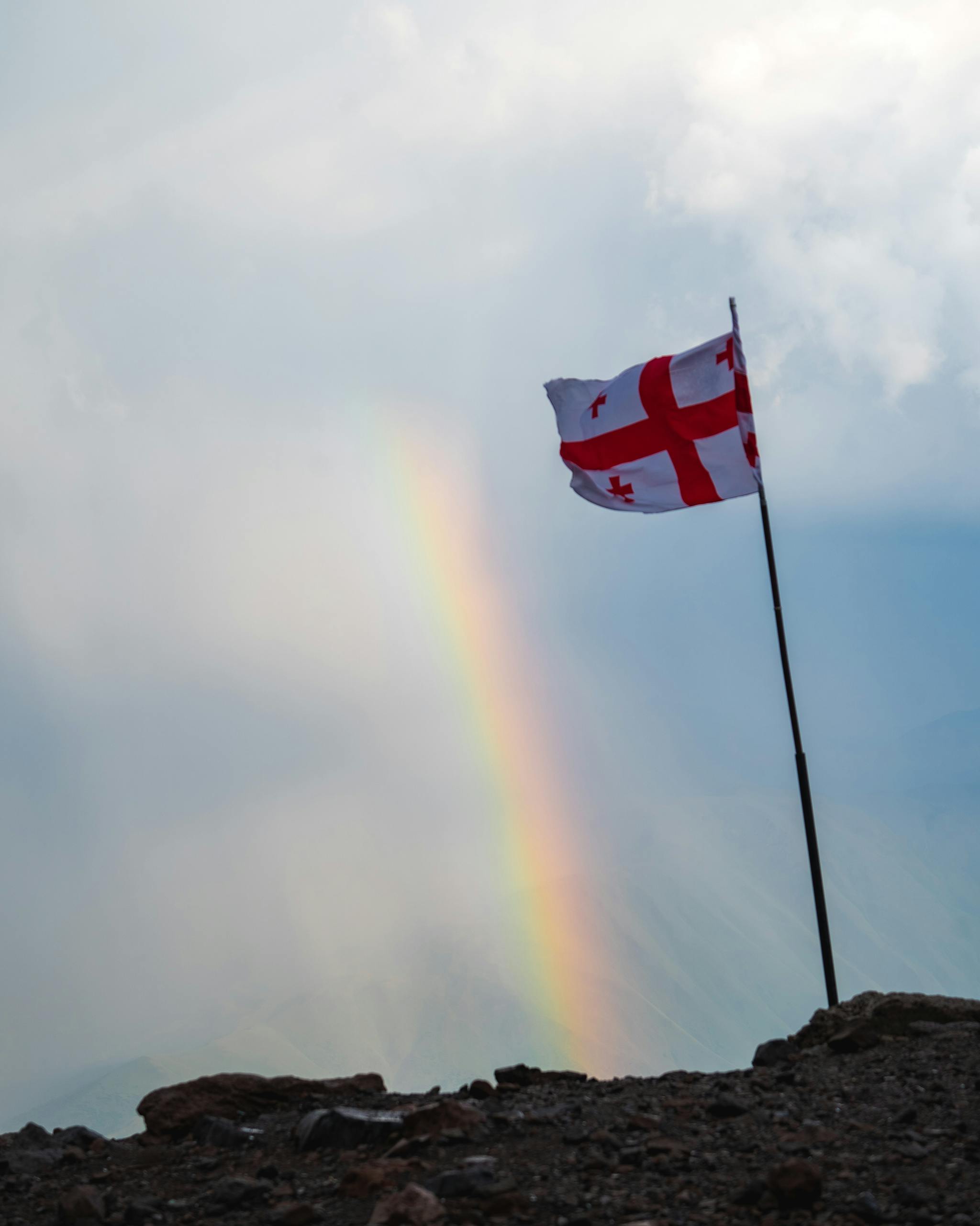Stunning view of the Georgian flag against a backdrop of a vibrant rainbow on a mountain peak.